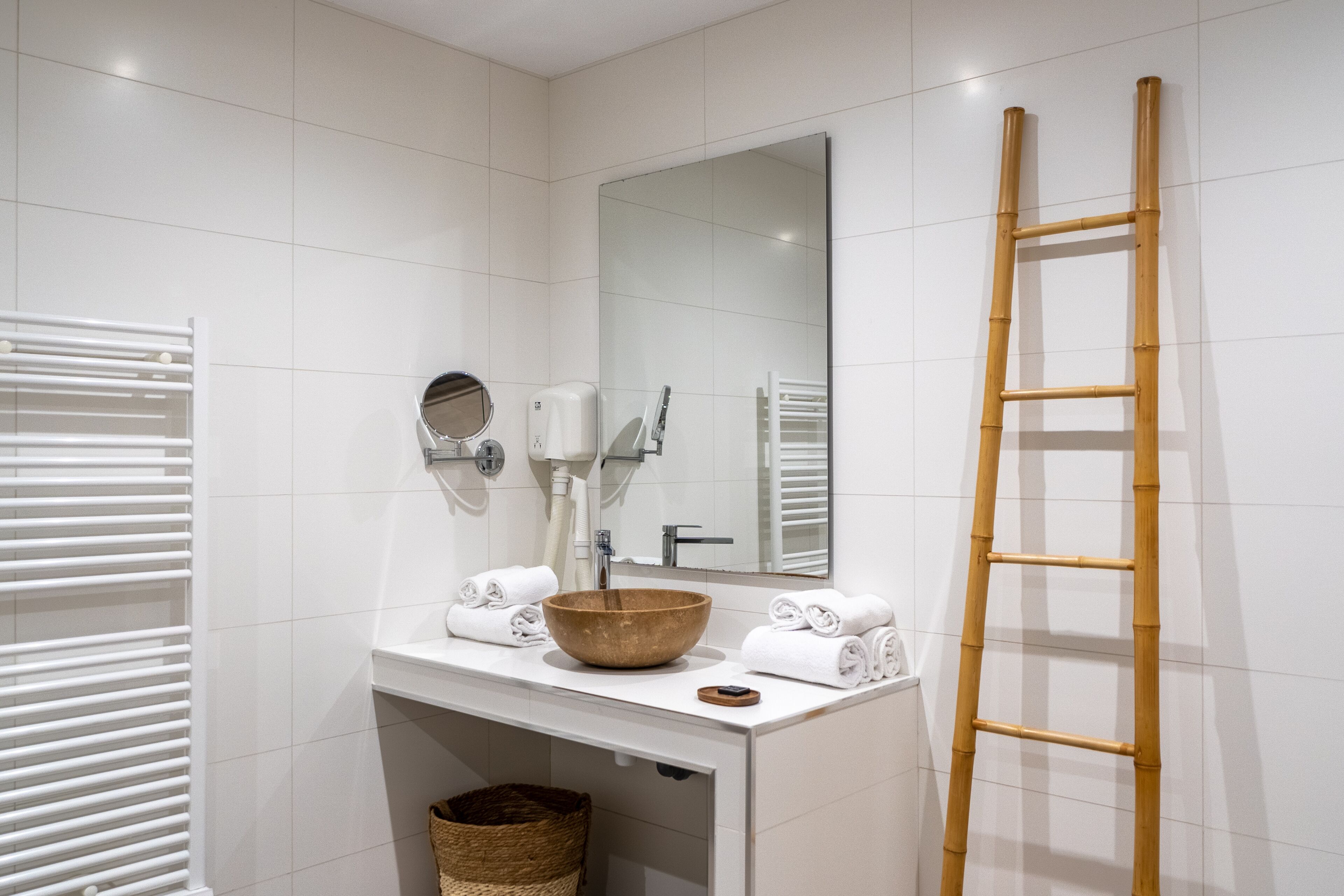 Ein Badezimmer mit weiß geflieste Wand, einem Waschbecken aus Holz, einem Spiegel, einem Handtuchhalter und einer Bambusleiter.