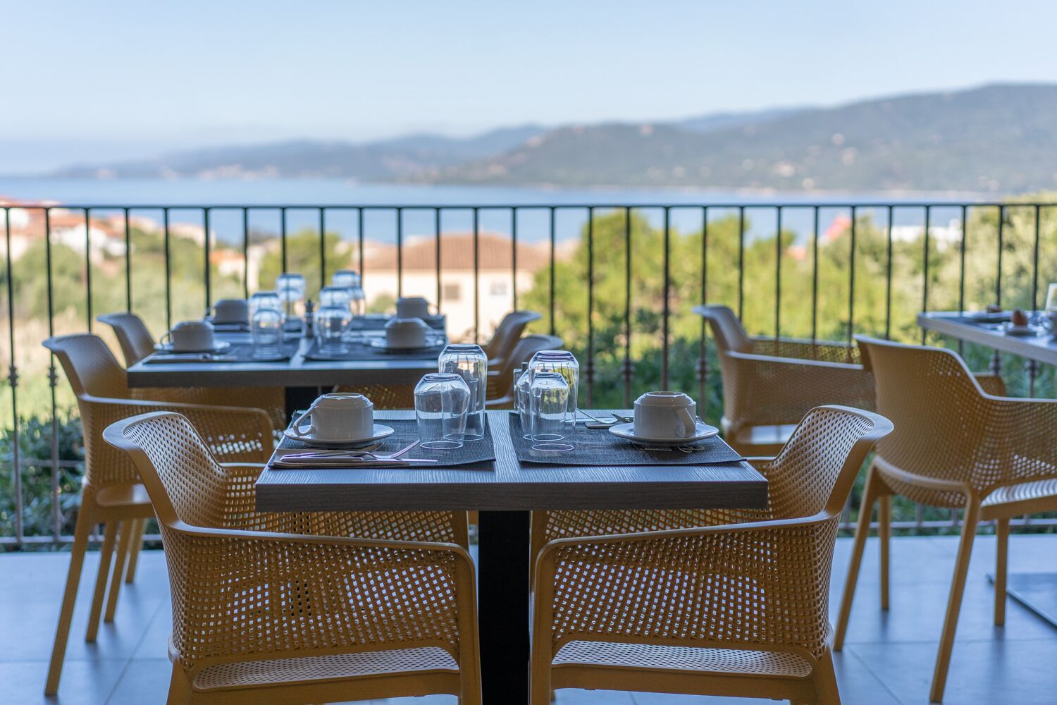 A dining area with tables set for four, outdoor chairs, and a scenic view of the sea and hills in the background.