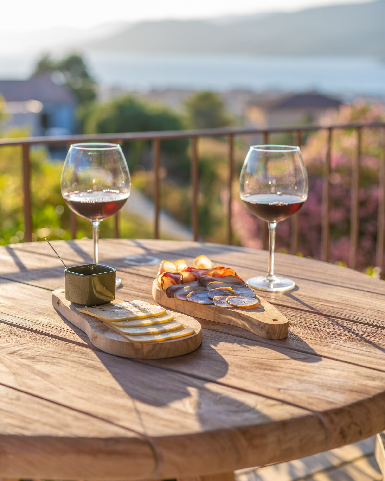 Una mesa de madera con dos copas de vino tinto, un cuenco pequeño y una tabla de madera con queso en rodajas y embutidos.
