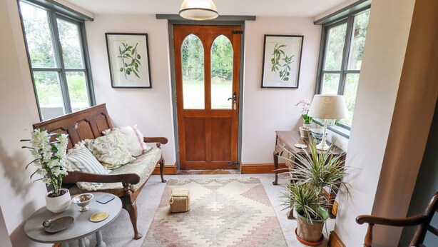 A cozy room with a wooden bench, a small table, and a sofa. There are framed botanical prints on the walls, a door with a wooden frame, and a window with a view of greenery. - The Waggon Shed (Nottingham)