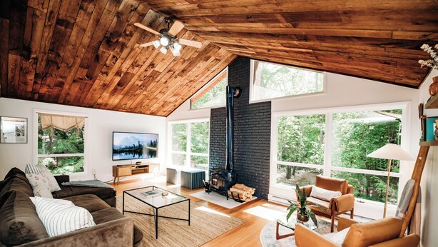 A spacious living room with a high ceiling, a fireplace, and large windows offering a view of the outdoors. - Riverfront FirePit! HotTub-Pool-Beaches-Gameroom (Gouldsboro)