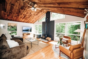 A spacious living room with a high ceiling, a fireplace, and large windows offering a view of the outdoors. - Riverfront FirePit! HotTub-Pool-Beaches-Gameroom (Gouldsboro)