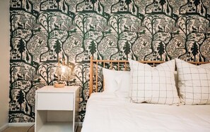 A bedroom with a patterned wallpaper, a wooden bed frame, a white bedside table with a lamp, and a white bedspread. - Riverfront FirePit! HotTub-Pool-Beaches-Gameroom (Gouldsboro)