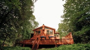 A wooden cabin with a deck, surrounded by dense forest. - Riverfront FirePit! HotTub-Pool-Beaches-Gameroom (Gouldsboro)