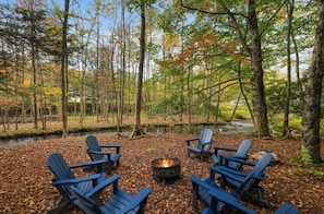 A forest setting with a fire pit and Adirondack chairs. - Riverfront FirePit! HotTub-Pool-Beaches-Gameroom (Gouldsboro)