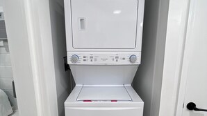 A compact, white laundry room with a front-loading washer and dryer, a small shelf above the dryer, and a door on the right. - Quaint cottage with a beautiful view. (Greenville)