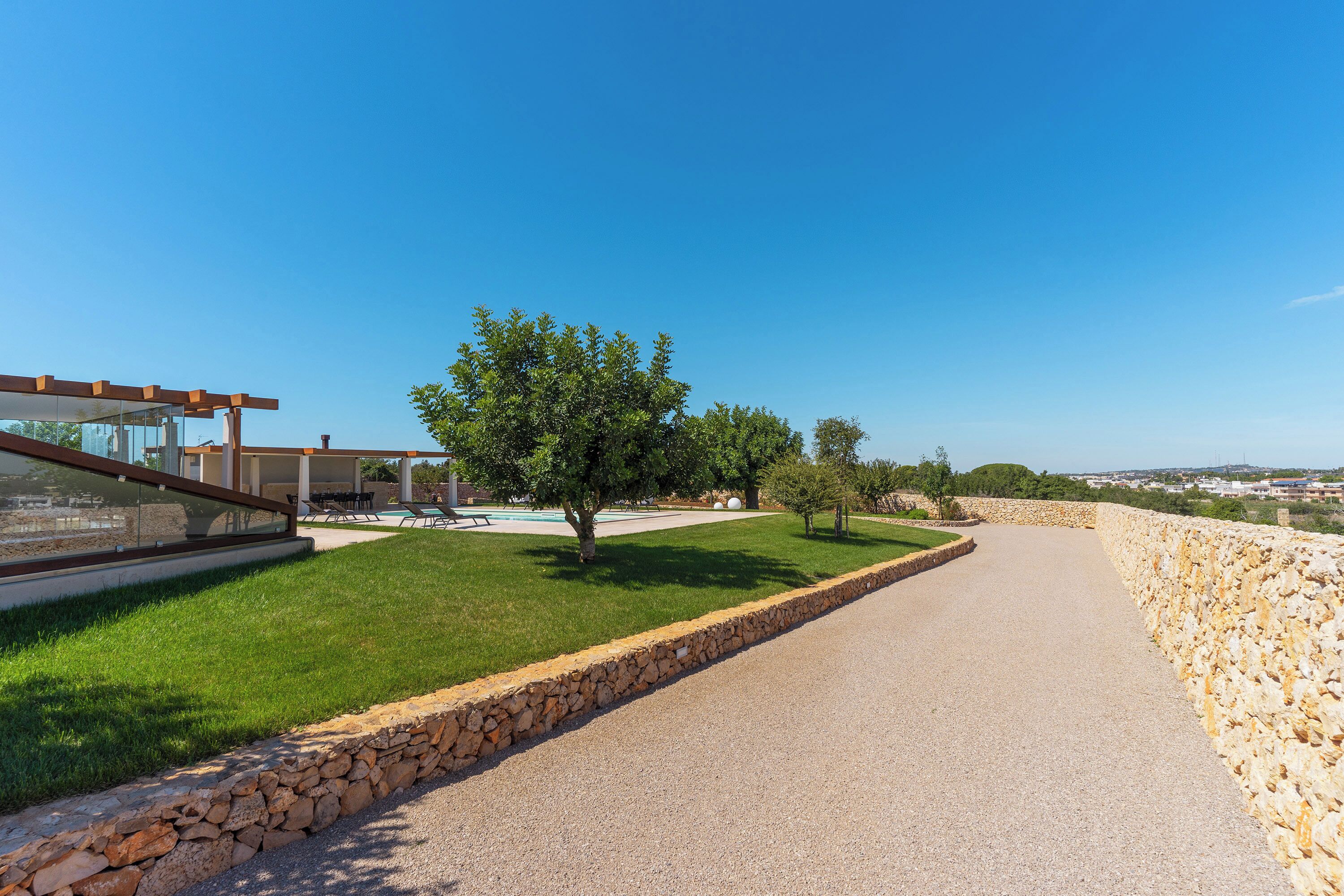 A paved pathway flanked by stone walls, a grassy area with a tree, and a modern building with a glass facade.