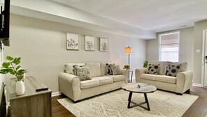 A living room with two beige sofas, a coffee table, a patterned rug, a floor lamp, and framed artwork on the wall. - Modern Townhome w/ Balcony • Walk to FUSE Stadium (Gastonia)
