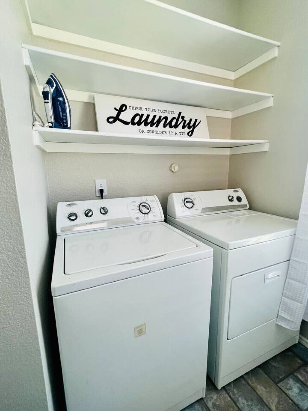 A laundry room with two white front-loading washing machines and a shelf above them with a sign that reads 'CHECK YOUR POCKETS Laundry OR CONSIDER IT A TIP'.