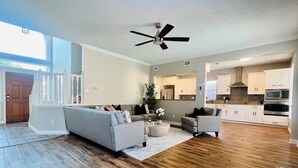 A spacious living room with hardwood flooring, a grey sofa, and a ceiling fan. - 6 Bedroom Home with Pool, Centrally Located (The Colony)