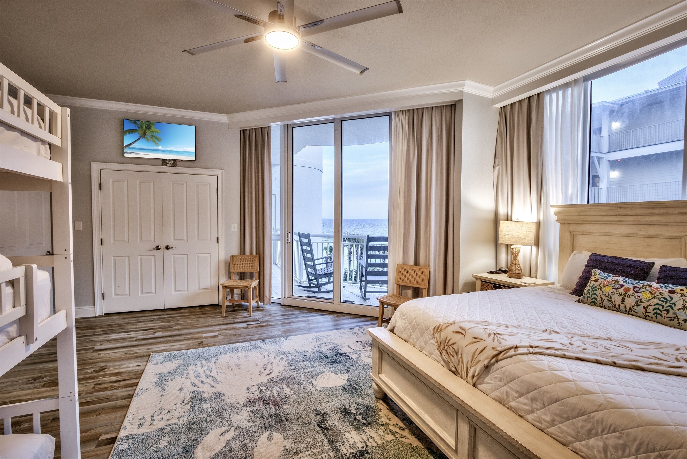 A hotel room with a bed, a ceiling fan, a TV on the wall, a sliding glass door to the balcony, and a wooden floor.