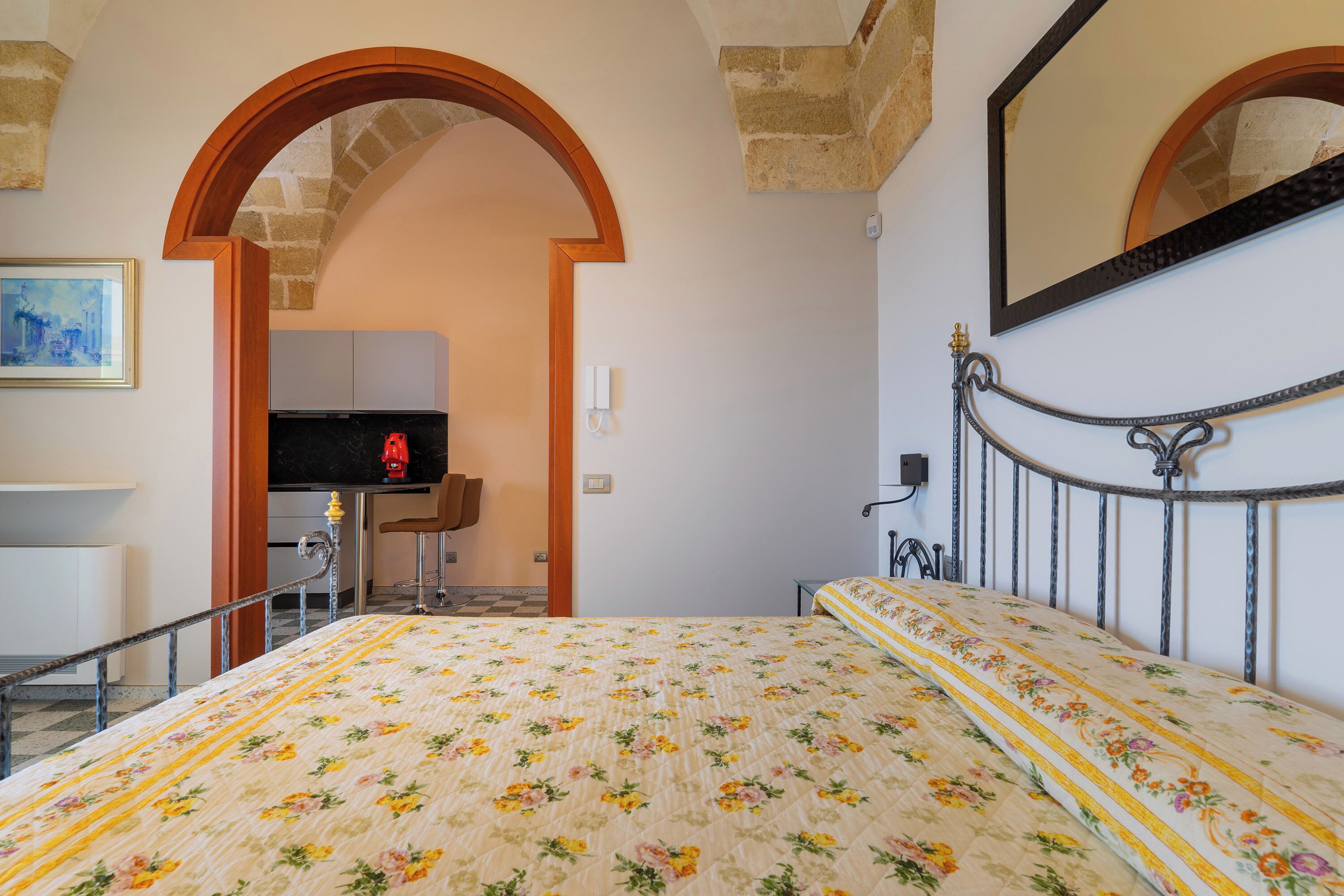 A bedroom with a floral bedspread, a metal-framed bed, a mirror, and a stone archway leading to a kitchenette.