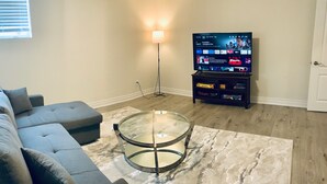 Ein Wohnzimmer mit grauem Sofa, einem Glastisch, einem Fernseher auf einem Schrank und einer Stehlampe.