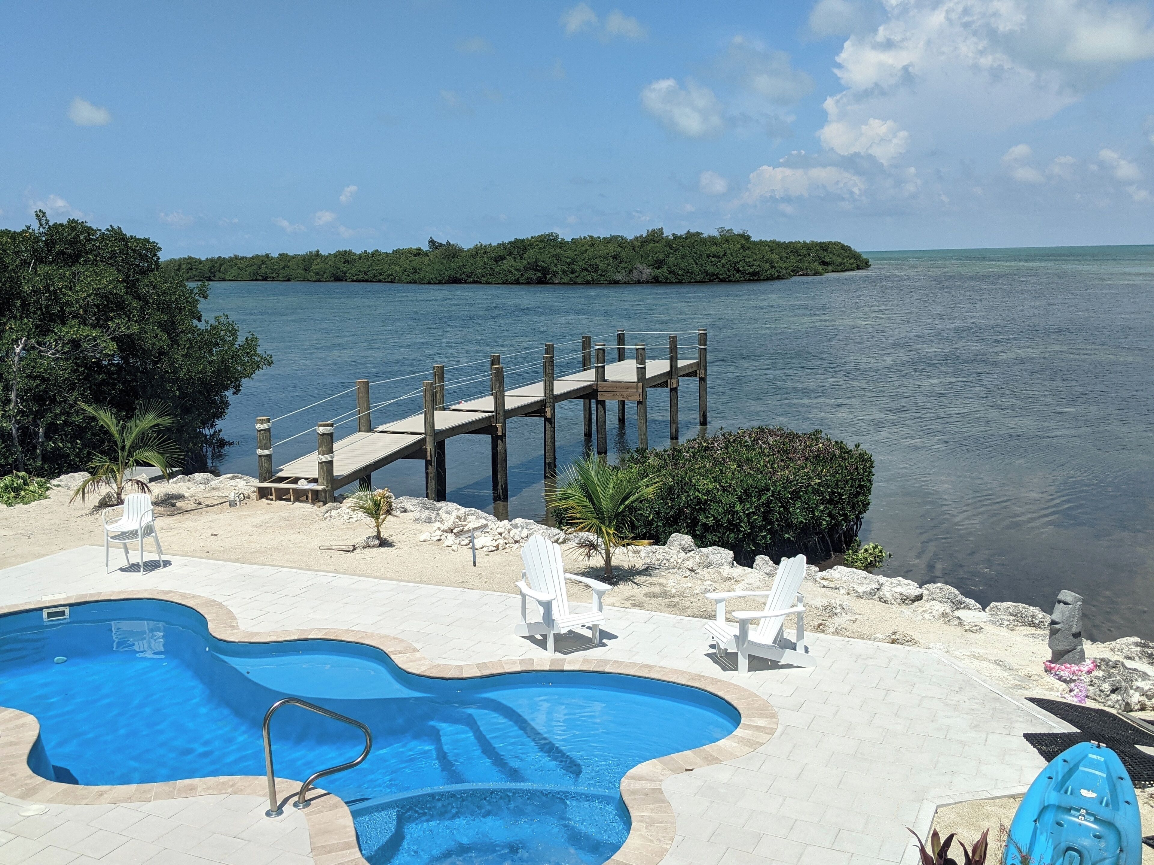 A swimming pool with a wooden deck extending over water, surrounded by lounge chairs and a small palm tree.