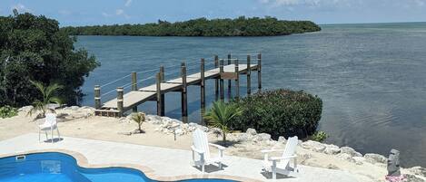 A swimming pool with a wooden deck extending over water, surrounded by lounge chairs and a small palm tree.