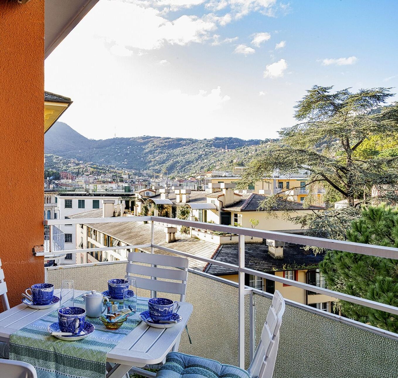 Ein Balkon mit einem für Tee gedeckten Tisch, mit Blick auf eine an einem Hügel gelegene Stadt mit Gebäuden und einem Berg im Hintergrund.