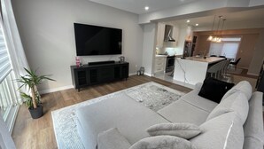 A modern living room with a large flat-screen TV, a grey sofa, and a dining area with a kitchen in the background. - Modern Townhouse, 5 Mins From all Amenities. (Guelph)
