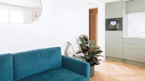 A modern living room with a teal sofa, a round mirror, a potted plant, and a kitchen area with a built-in oven and cabinets. - Sleek 1BD Flat 5 Mins From City Centre - Brighton (Brighton)
