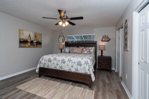A bedroom with a bed, a ceiling fan, a nightstand, a lamp, a window, and a door.