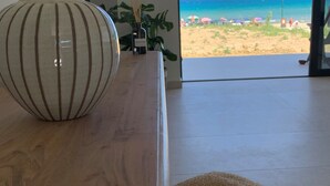 Ein Zimmer mit einem Holztisch, einer großen Vase und einem Korbsitzhocker. Ein Fernseher ist an der Wand befestigt, und durch eine geöffnete Tür ist der Blick auf einen Strand möglich.