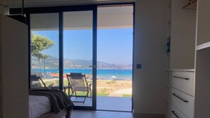 Ein Schlafzimmer mit einem Bett, einem Sessel und Blick auf Strand und Meer durch eine Schiebetür.