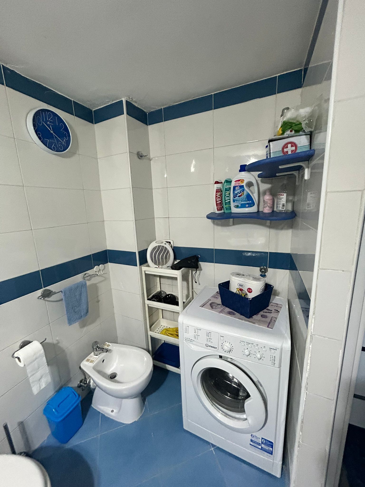 A compact bathroom with a toilet, bidet, and washing machine. There are shelves with toiletries and a clock on the wall.