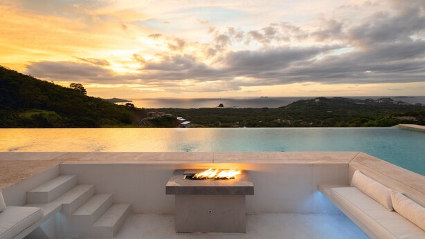 A infinity pool with a fire feature, white outdoor seating, and a scenic view of a coastline and hills during sunset. - 5BR home|Mar Vista|Ocean View, Pool, Deck|Fire Pit – Casa Ayla A (Playa Flamingo)