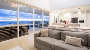 A modern living room with a grey sofa, a dining area with a white table, and a kitchen with white cabinets and pendant lights. - 5BR home|Mar Vista|Ocean View, Pool, Deck|Fire Pit – Casa Ayla A (Playa Flamingo)
