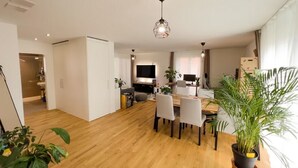 Un salon moderne avec un plancher en bois, un coin repas comprenant une table et des chaises, un téléviseur et une grande fenêtre avec des rideaux.