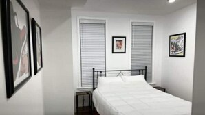A bedroom with a bed, two bedside tables, a window with blinds, and framed pictures on the wall. - City Arts Apartment (Philadelphia)