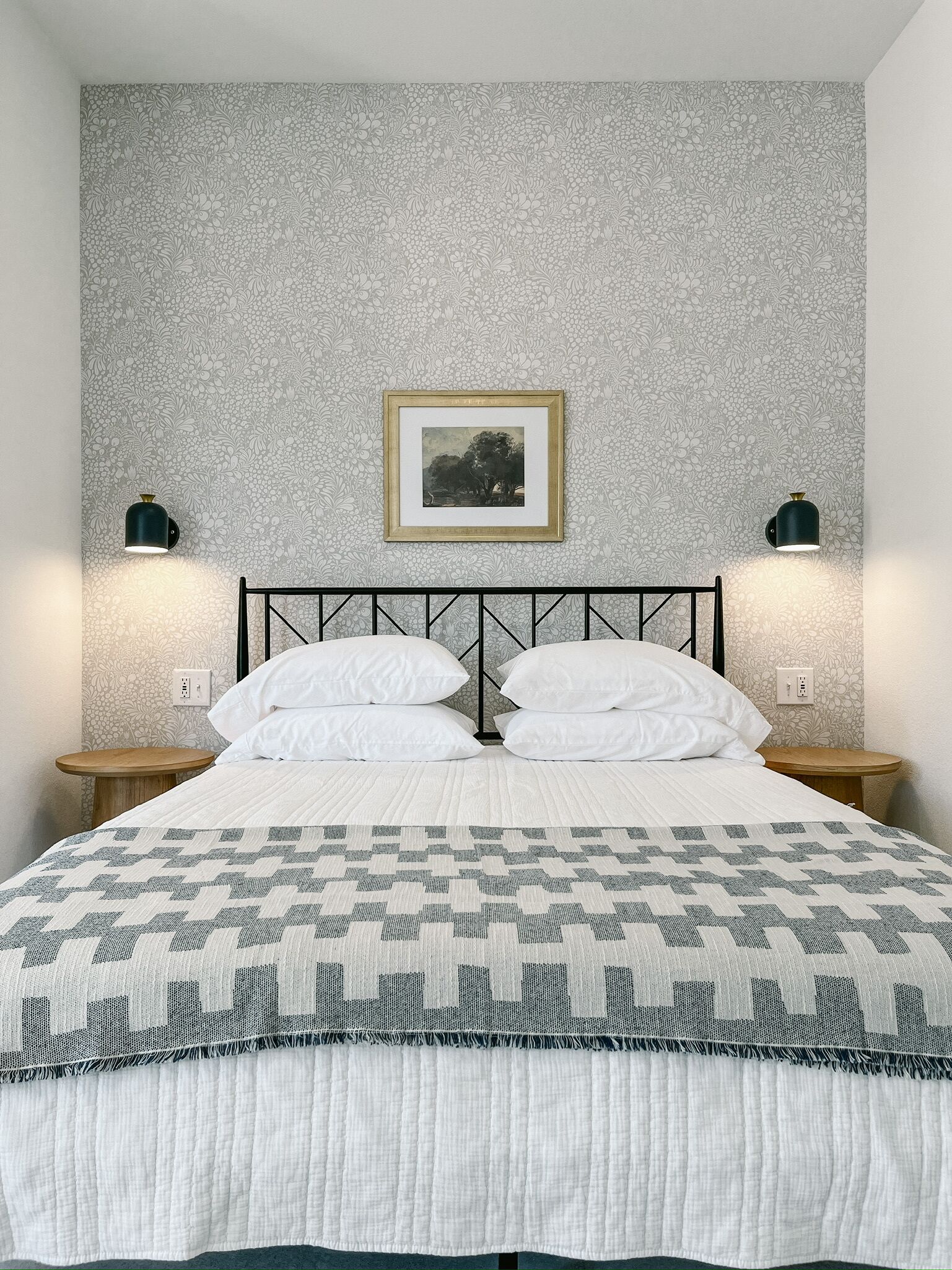 A neatly made bed with a patterned bedspread, a framed picture on the wall, and two wall-mounted lamps.