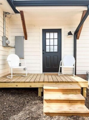Une terrasse en bois avec deux chaises blanches, une porte noire et un petit banc en bois.