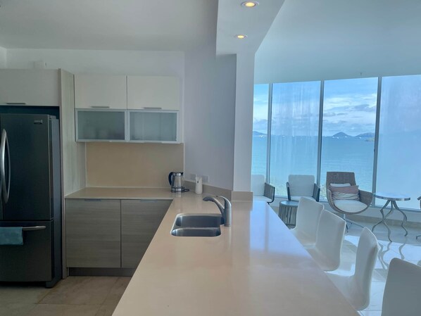 A modern kitchen with a stainless steel refrigerator, a sink, and a coffee maker. There are white cabinets and a large window offering a view of the sea.