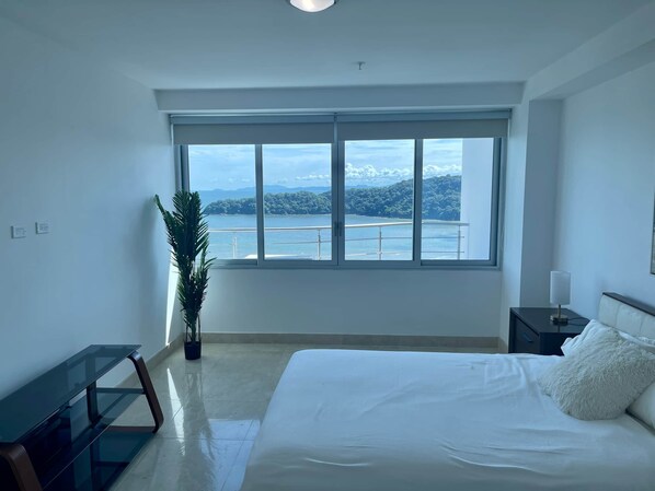 Ein Schlafzimmer mit großem Fenster, einem Bett, einem Nachttisch und einer Pflanze.
