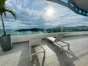 A balcony with two white wicker chairs, a potted plant, and a glass railing overlooking a scenic view.