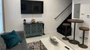 A living room with a grey sofa, teal cushions, a patterned rug, a coffee table, a flat-screen TV, a blue cabinet, and a staircase with a black railing.