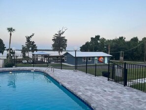 A swimming pool with a paved deck, surrounded by a fence and a building with a '76' sign.