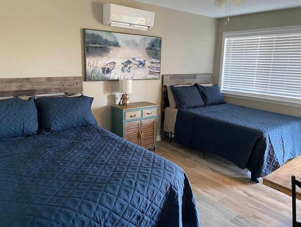 A bedroom with two beds, a wooden headboard, a painting of boats, a small dresser, and a window with blinds.