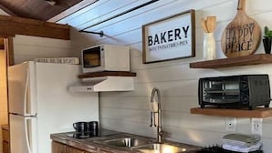 Cozinha com armários de madeira, pia de aço inoxidável, micro-ondas e torradeira. Há um letreiro com a palavra 'BAKERY' e outro que diz 'ESTE É O NOSSO LUGAR FELIZ'.