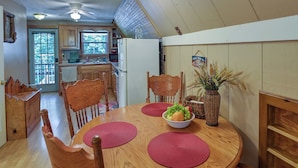 Un coin repas avec une table en bois, des sets de table rouges et un saladier rempli de fruits. On y trouve des chaises en bois et un ventilateur de plafond. On aperçoit une cuisine à l’arrière-plan, avec des meubles de cuisine en bois et un réfrigérateur.