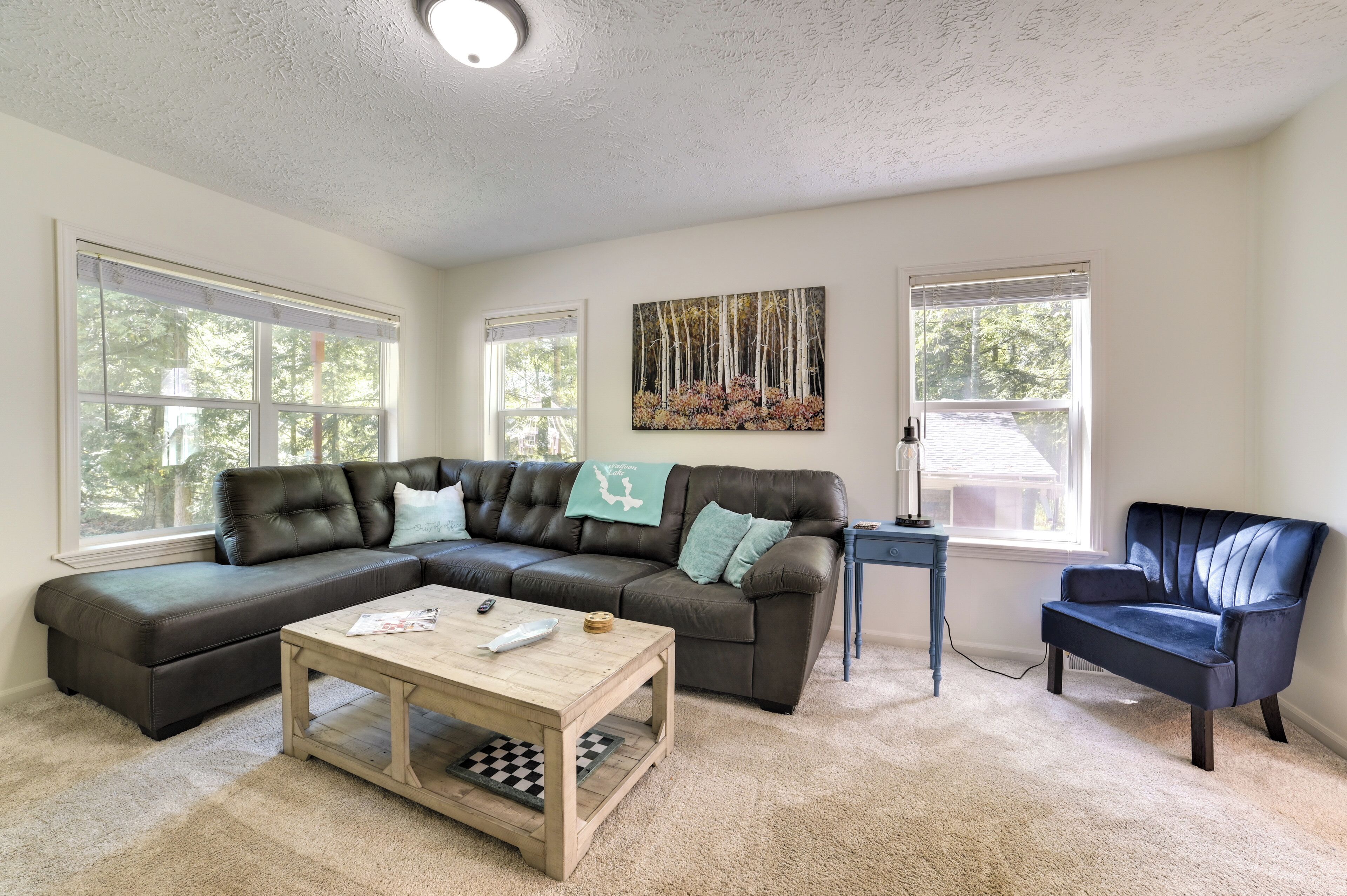 A living room with a sectional sofa, a coffee table, and a chair. There are two windows with blinds, a painting on the wall, and a lamp on a side table.