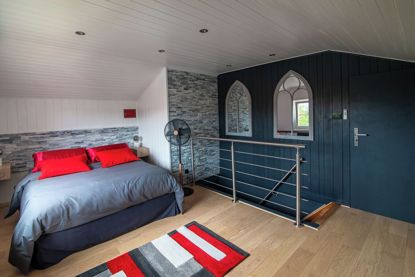 A bedroom with a bed, a standing fan, a window with a view, and a dark door.
