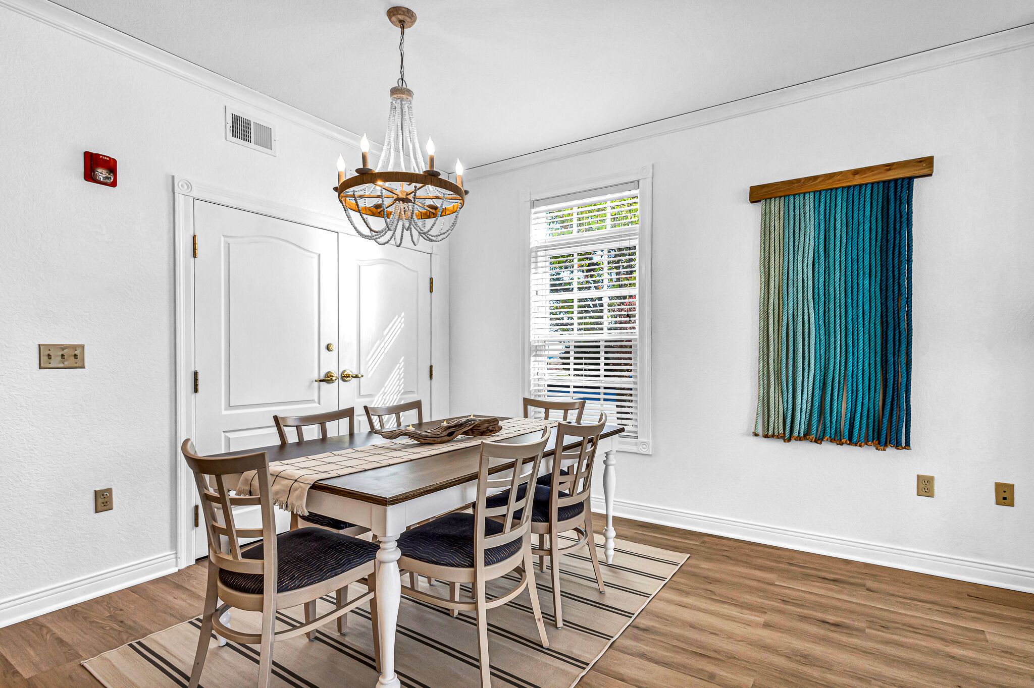 Un comedor con una mesa y sillas de madera, un candelabro, una ventana con persianas y una cortina azul.