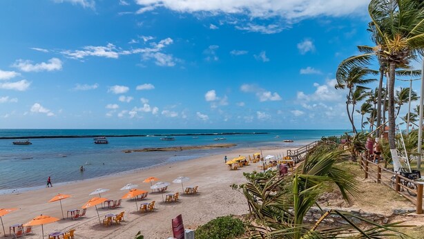 A beach with umbrellas and chairs, palm trees, and boats in the water. - Caravelas (Marechal Deodoro)