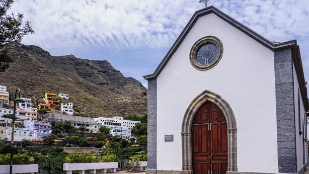 A white church with a wooden door and a clock above it, surrounded by a paved area and a hillside with buildings in the background. - Casa Yeyo, Lomo Bermejo (Santa Cruz de Tenerife)