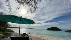 Une terrasse en bois, agrémentée d’un parasol vert, qui offre une vue sur une plage aux eaux calmes et une petite île au loin.