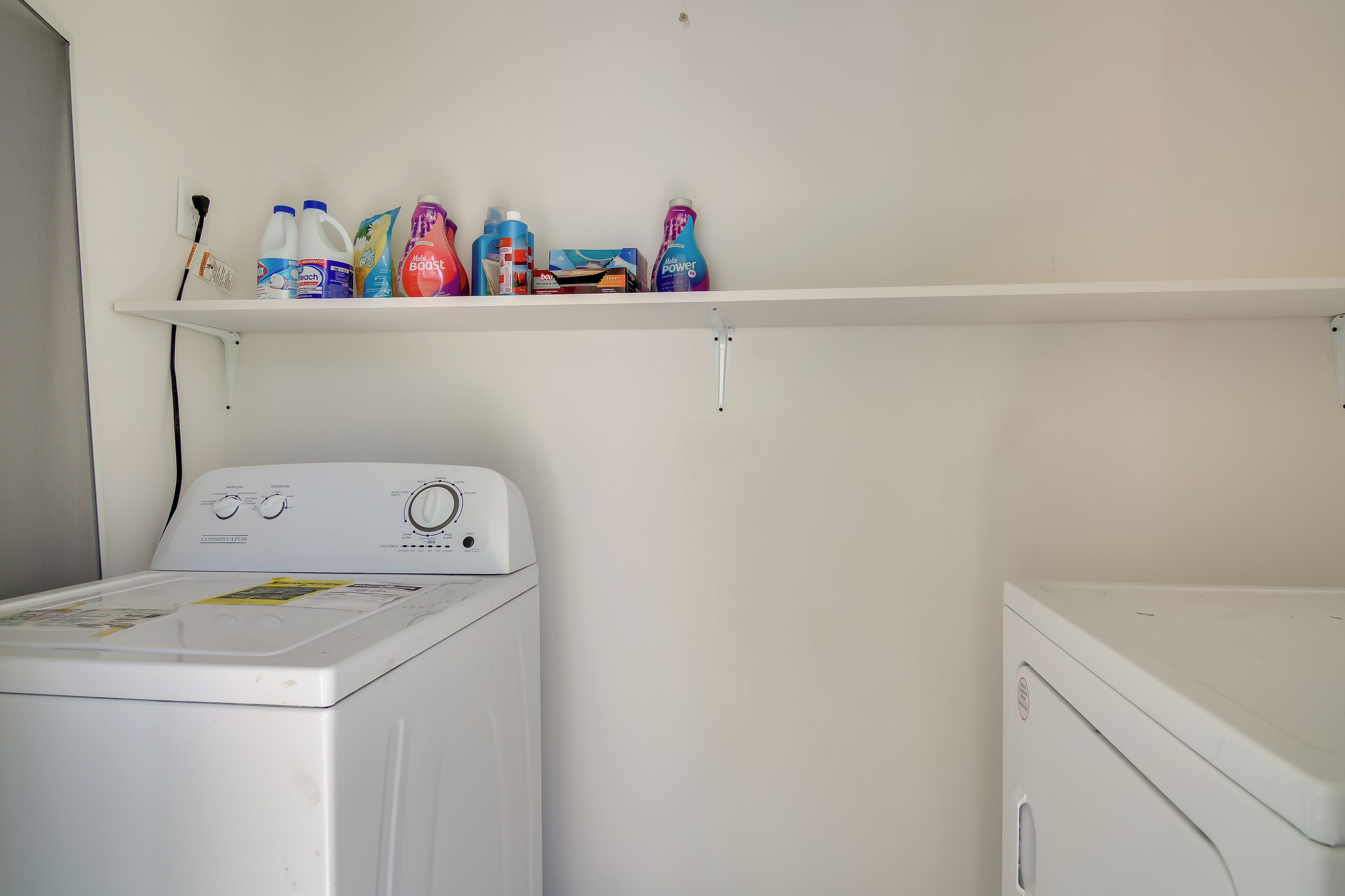 A laundry area with a washing machine and a dryer, a shelf with various cleaning products, and a power outlet.