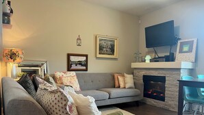 Una sala de estar con un sofá gris, una chimenea, un televisor montado y un cuadro en la pared.