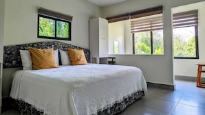 A modern bedroom with a large bed, two windows with blinds, and a view of greenery outside.