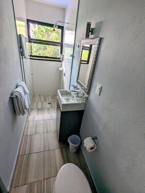 Un baño estrecho con inodoro, lavamanos y espejo. Hay una ventana que deja entrar luz natural y ofrece vistas a zonas verdes.